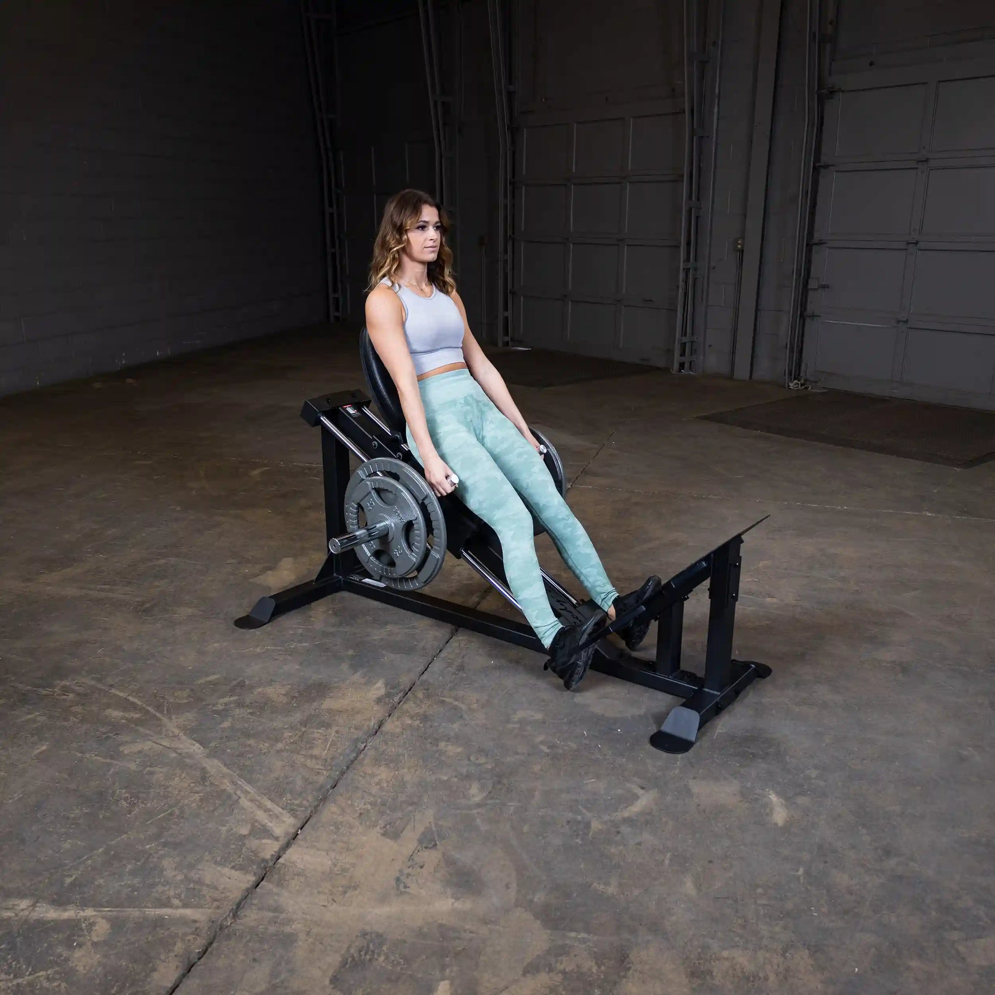 Woman using a Rogue leg press machine in a gym, wearing green leggings and a gray top.