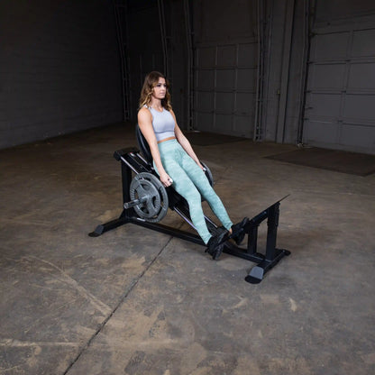 Woman using a Rogue leg press machine in a gym, wearing green leggings and a gray top.