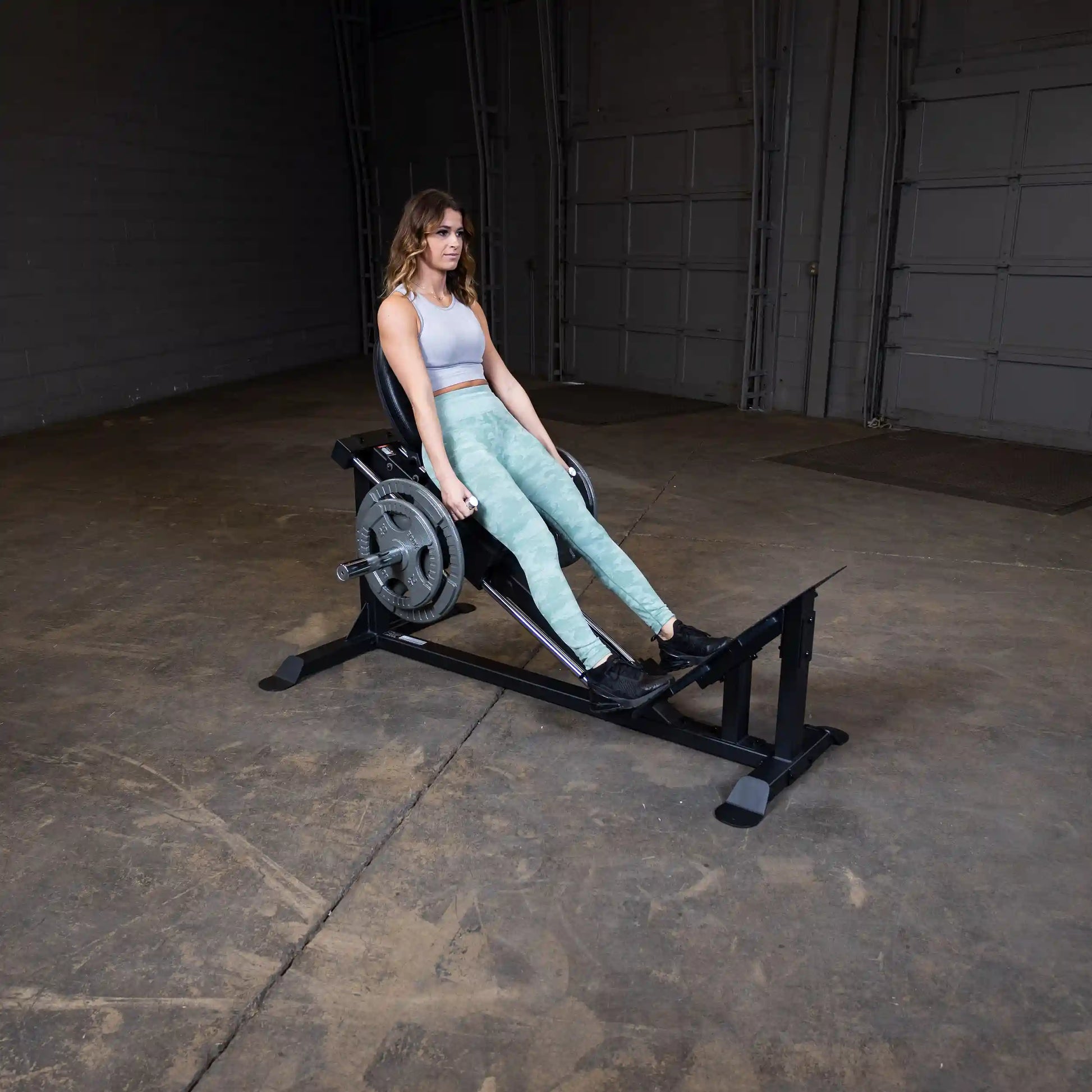 Woman using a leg press machine in a gym, wearing green leggings and a gray top.