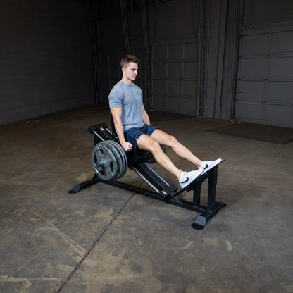 Man using a leg press machine in a gym, wearing a gray shirt and blue shorts.