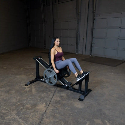 Woman exercising on black leg press machine in a gym, wearing purple top and gray leggings.