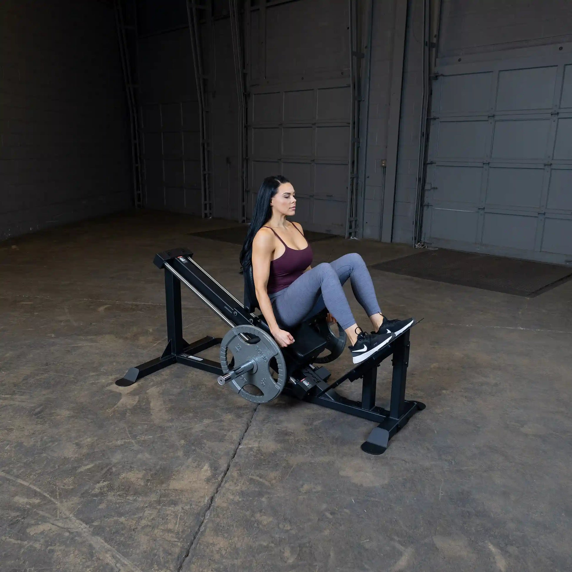 Woman using a leg press machine in a gym, wearing a maroon tank top and gray leggings.