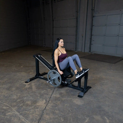 Woman using a leg press machine in a gym, wearing a maroon tank top and gray leggings.