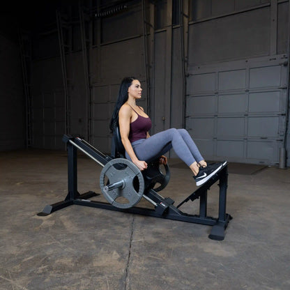 Woman using leg press machine in gym wearing purple top and gray leggings.