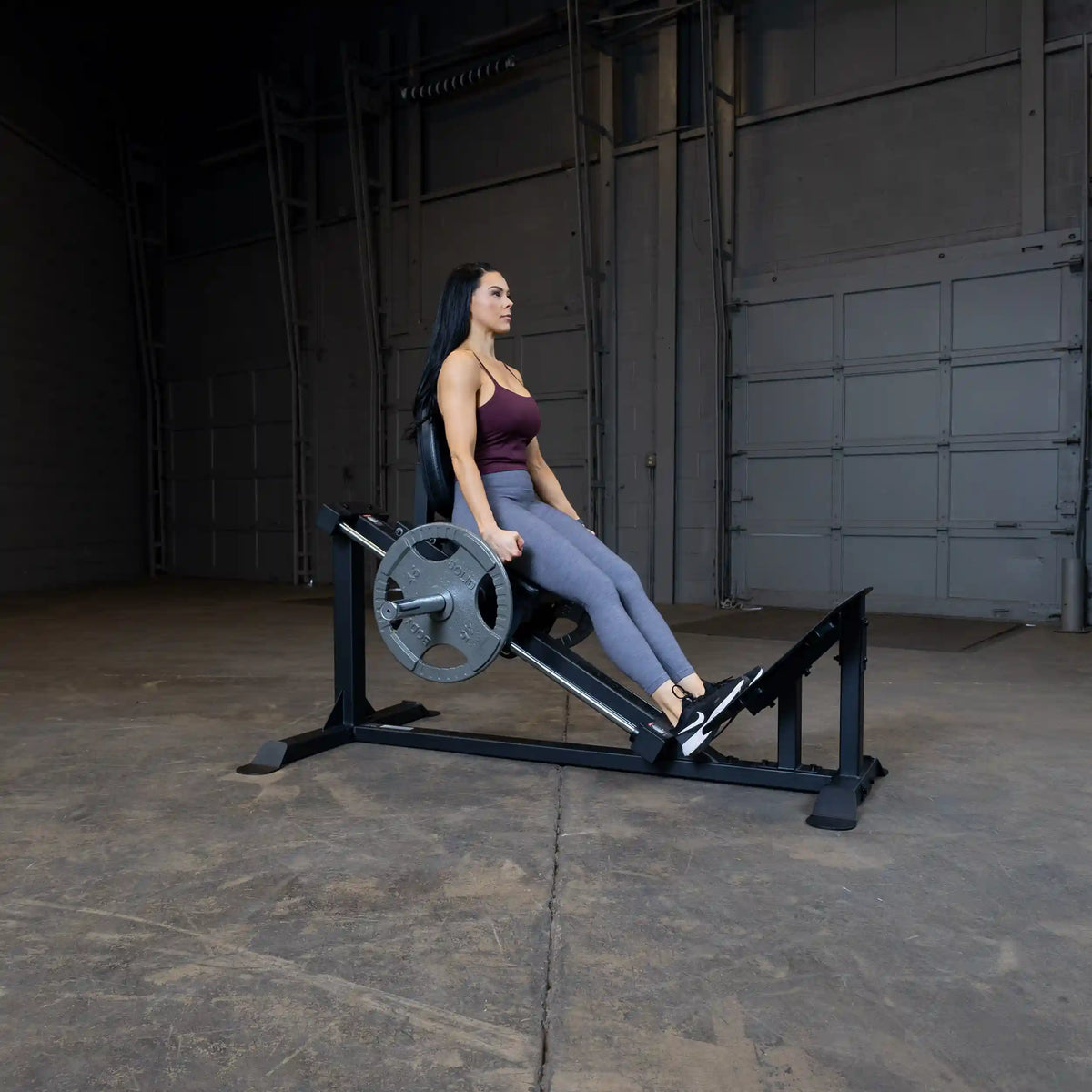 Woman using leg press machine in gym, wearing maroon top and gray leggings.
