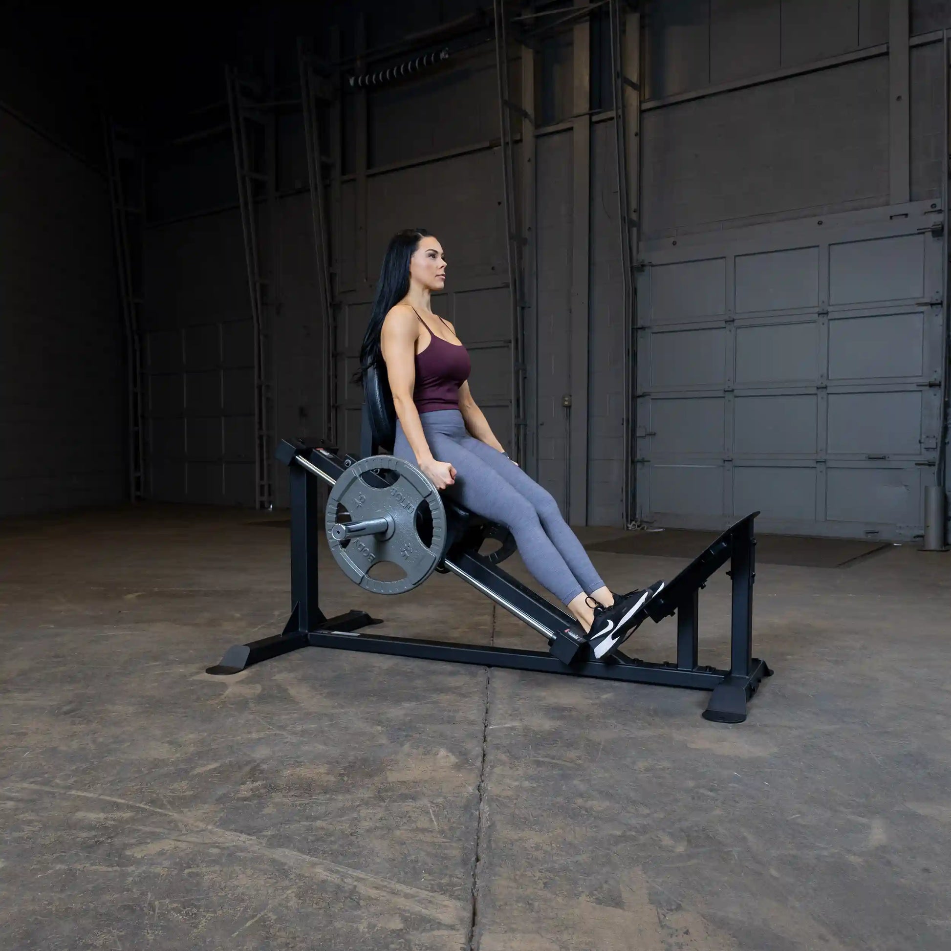 Woman using leg press machine in gym, wearing maroon top and gray leggings.