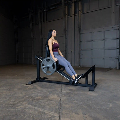 Woman using leg press machine in gym, wearing maroon top and gray leggings.