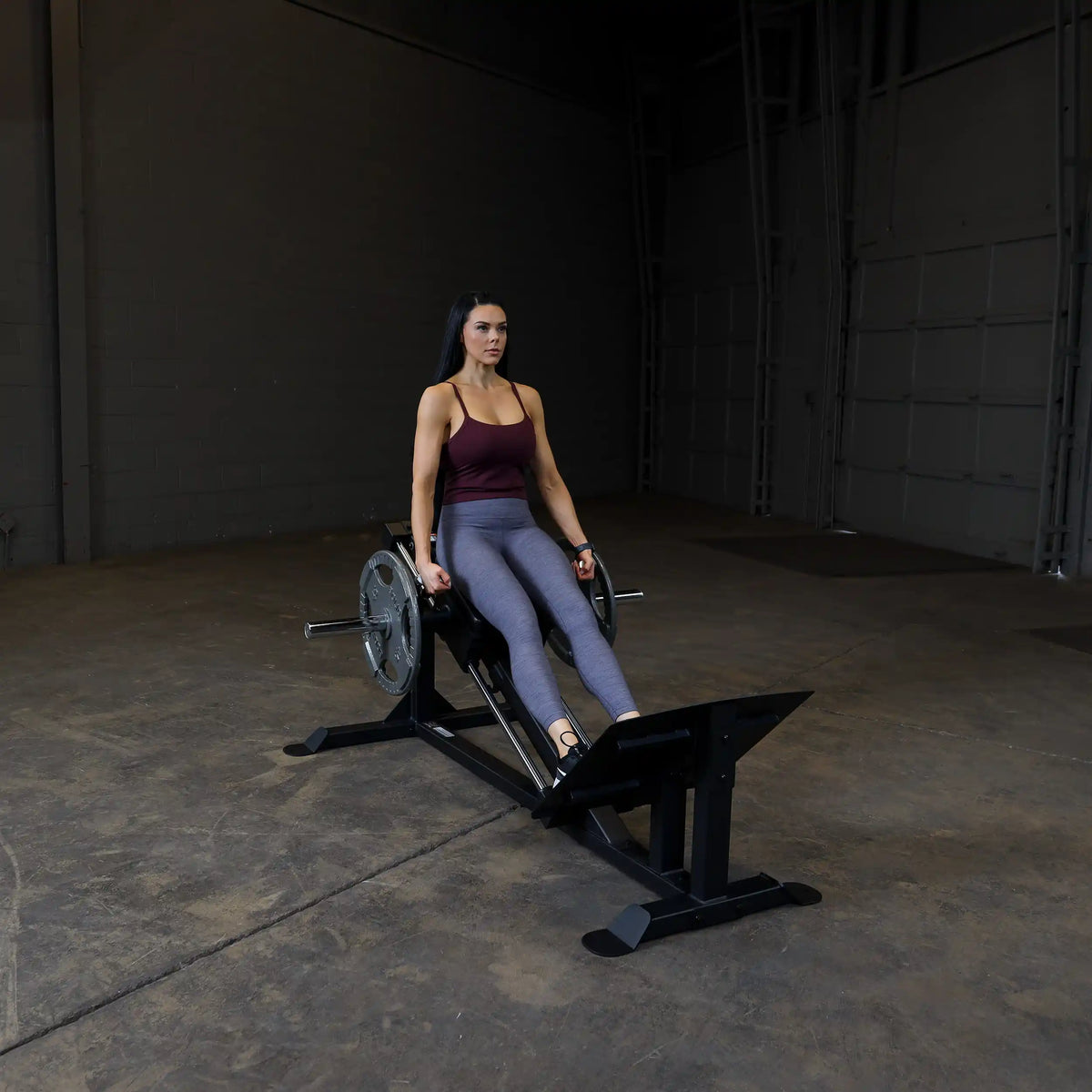 Woman using a black leg press machine in a gym, wearing gray leggings and a maroon top.