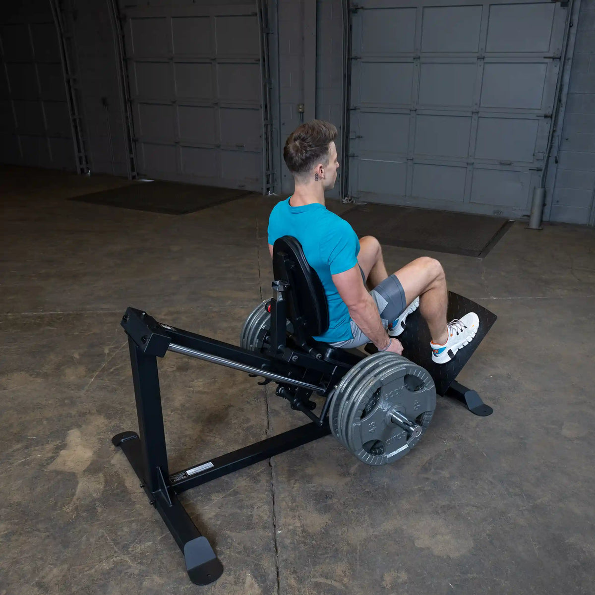 Man using a leg press machine in a gym, wearing a blue shirt and gray shorts, focusing on leg workout.