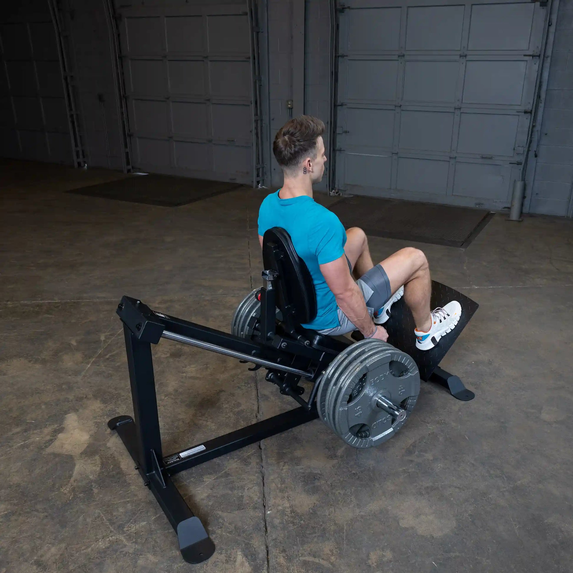 Man using a leg press machine in a gym, wearing a blue shirt and gray shorts, focusing on leg workout.