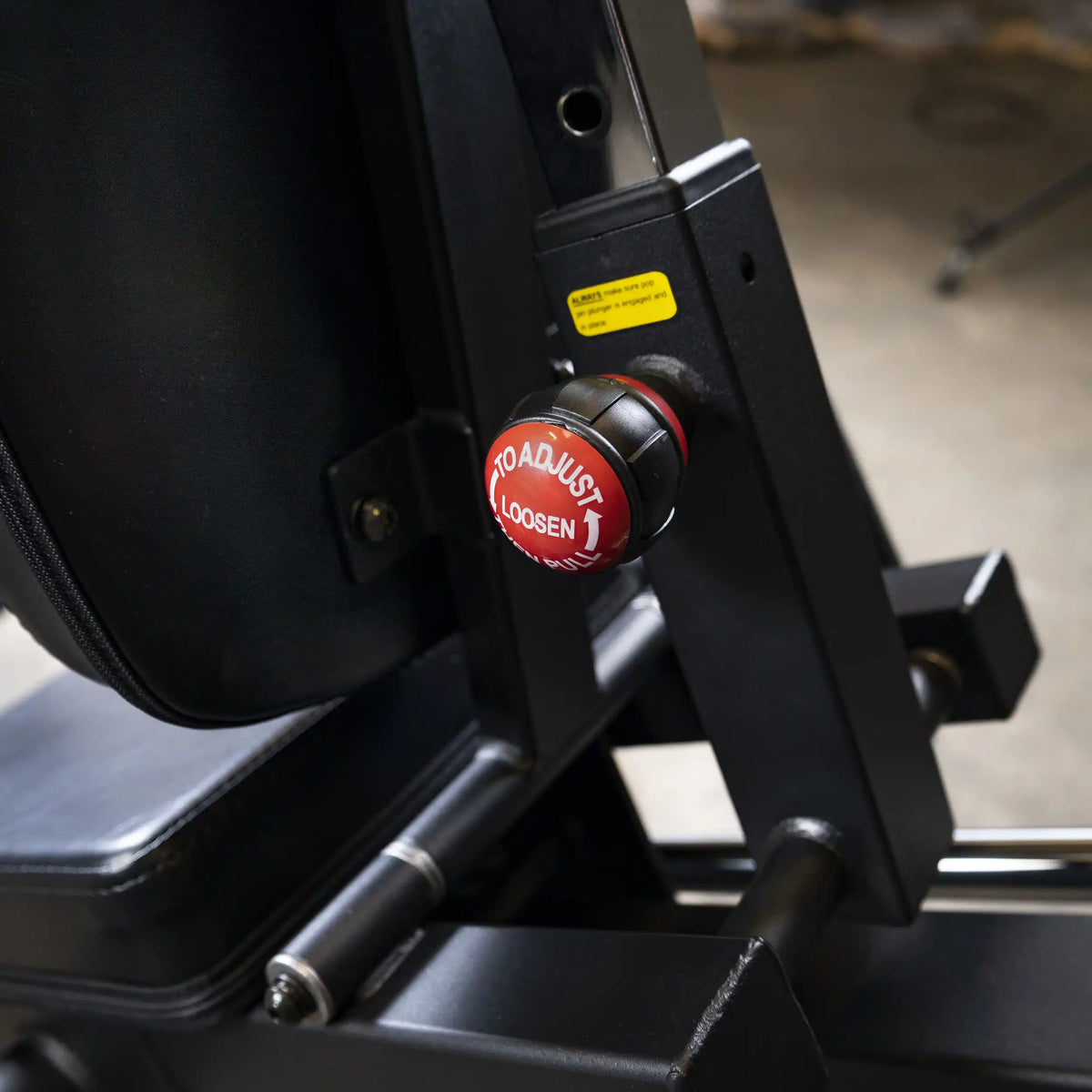 Adjustable gym equipment knob with red To Adjust Loosen label, featuring black metal frame.