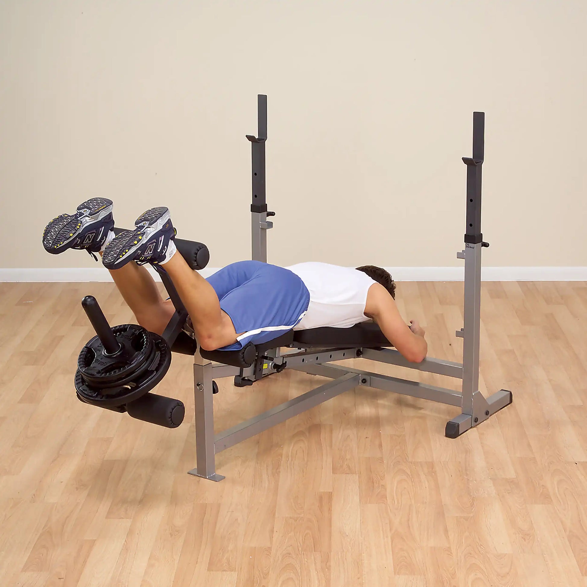 Man exercising on a weight bench with leg curl attachment, wearing blue shorts and sneakers.