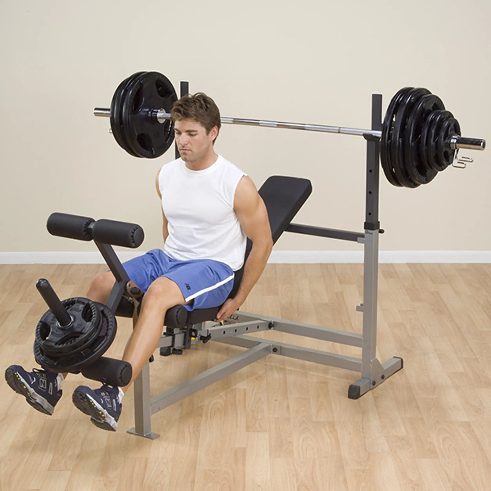 Man using a weight bench for leg exercises, wearing blue shorts and a white tank top, in a home gym setting.