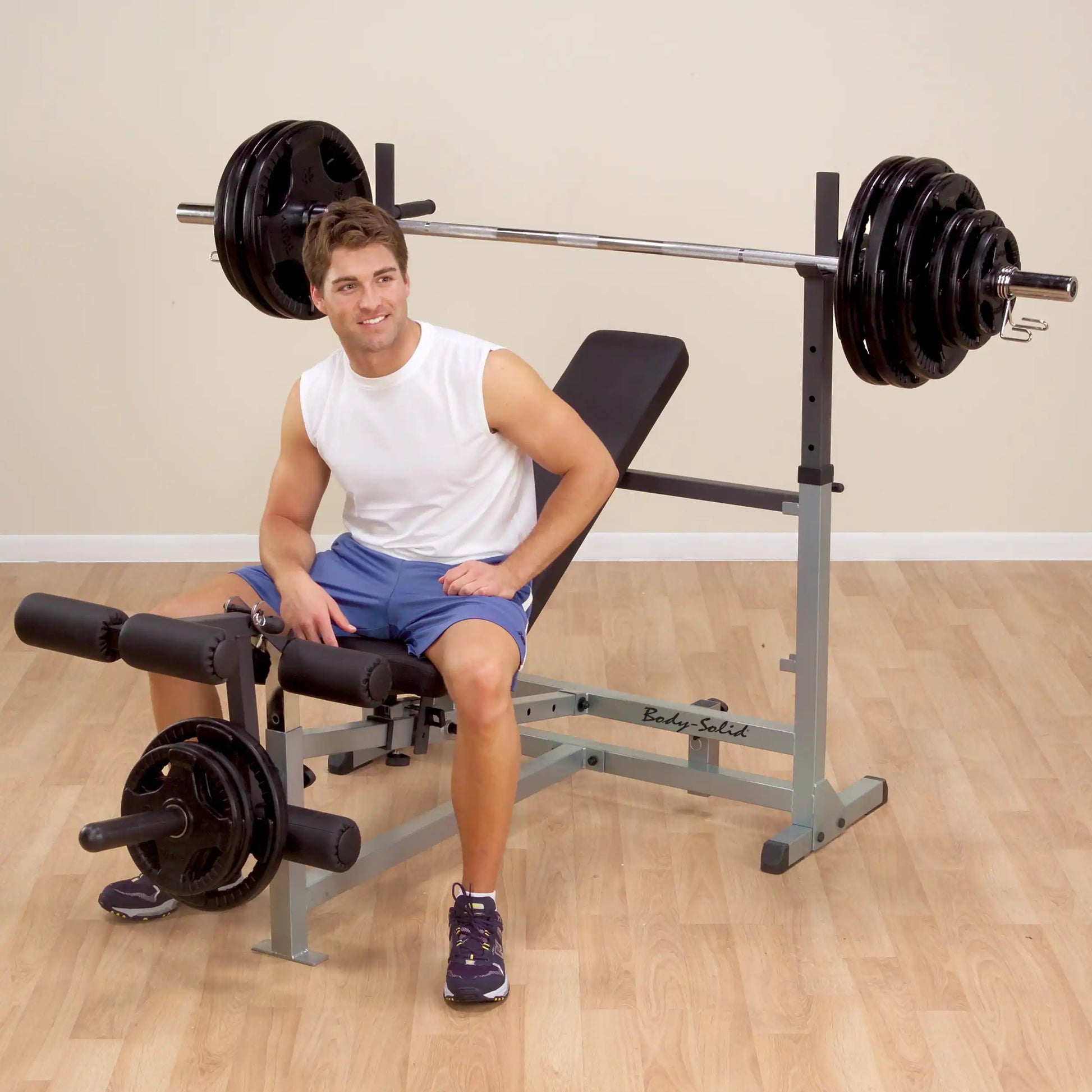 Man sitting on Body-Solid weight bench with barbell, wearing white tank top and blue shorts, in home gym.