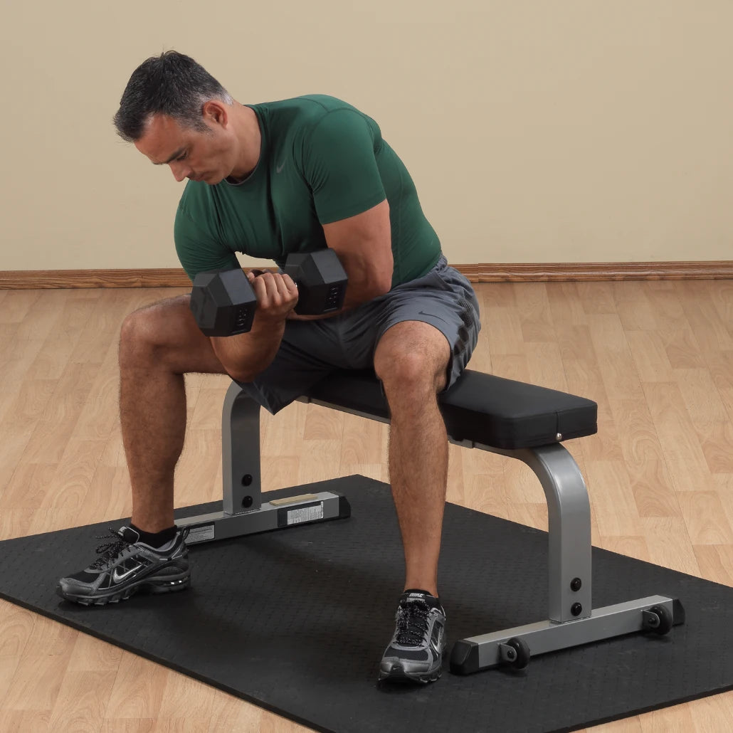 Man in green shirt performing dumbbell curls on a flat weight bench, emphasizing arm workout.