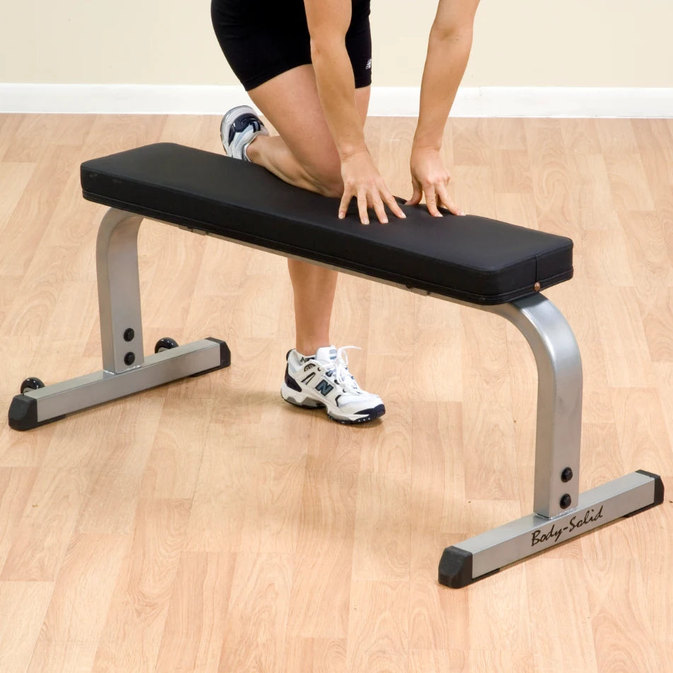 Woman stretching on Body-Solid flat bench with black cushion and silver frame on wooden floor.