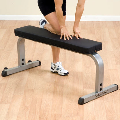 Woman stretching on Body-Solid flat bench with black cushion and silver frame on wooden floor.