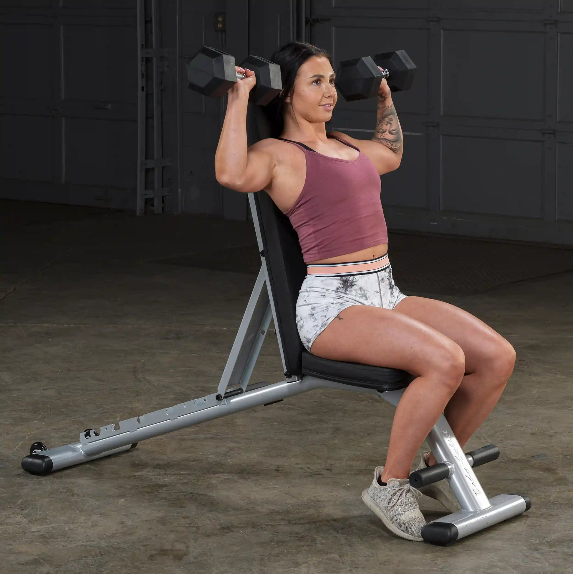 Woman performing shoulder press on Bowflex bench with dumbbells in gym.