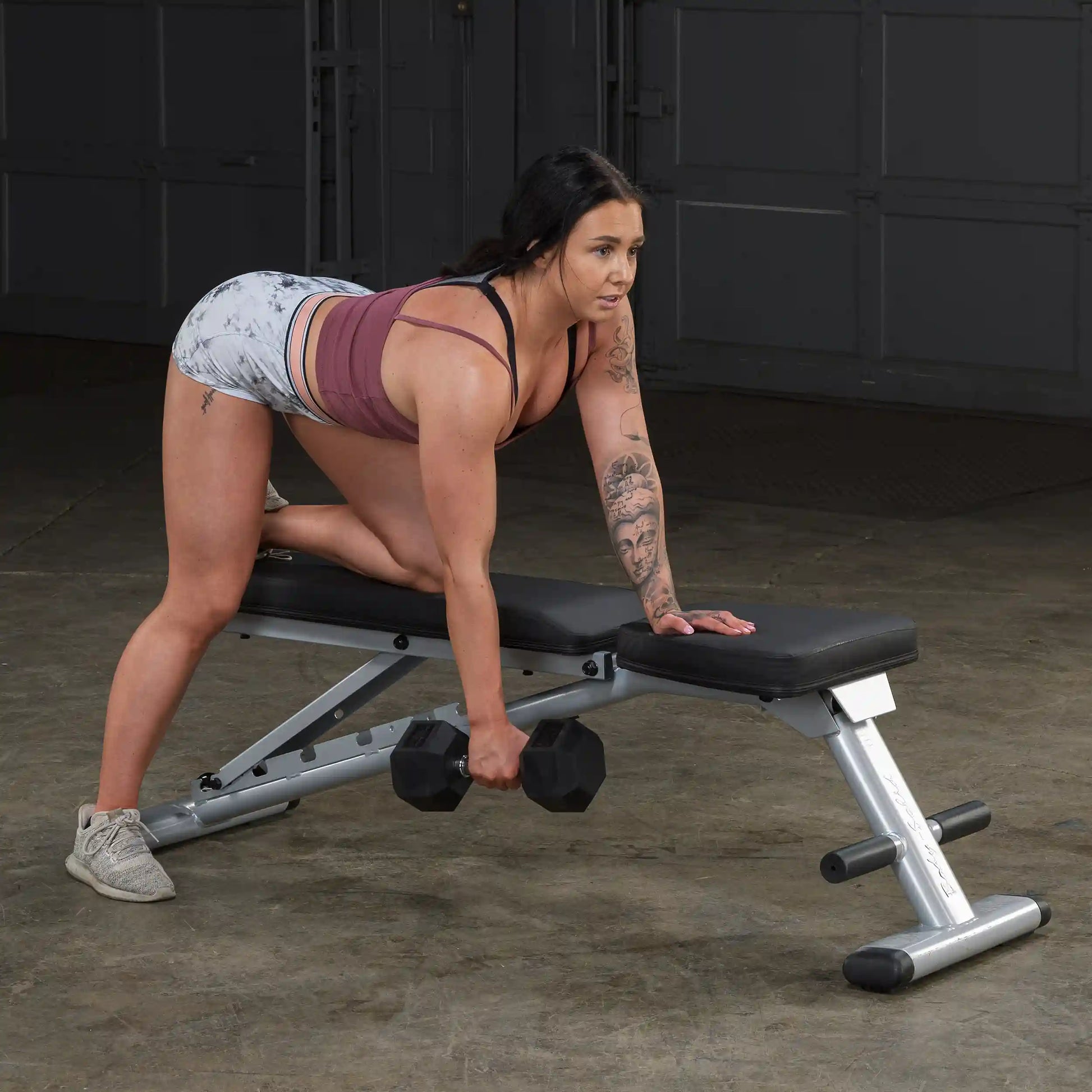 Woman performing dumbbell row on black workout bench, wearing maroon top and patterned shorts.