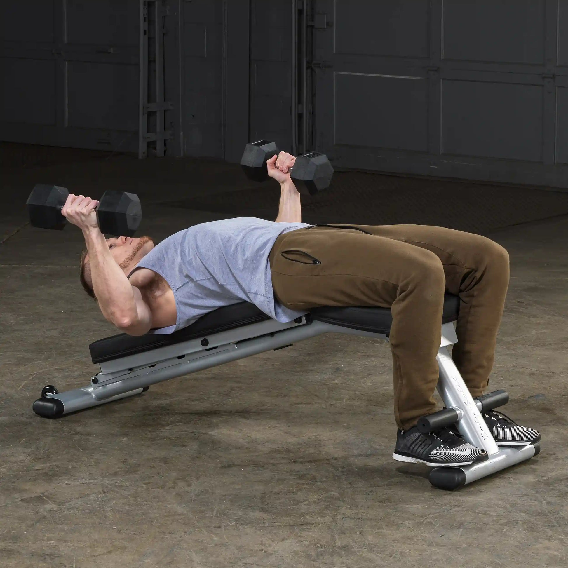 Man performing dumbbell bench press on an incline bench wearing brown pants in a gym.