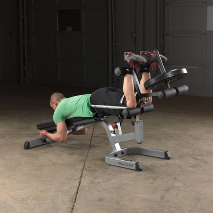 Man using Body-Solid leg curl machine with weight plates in a garage gym.