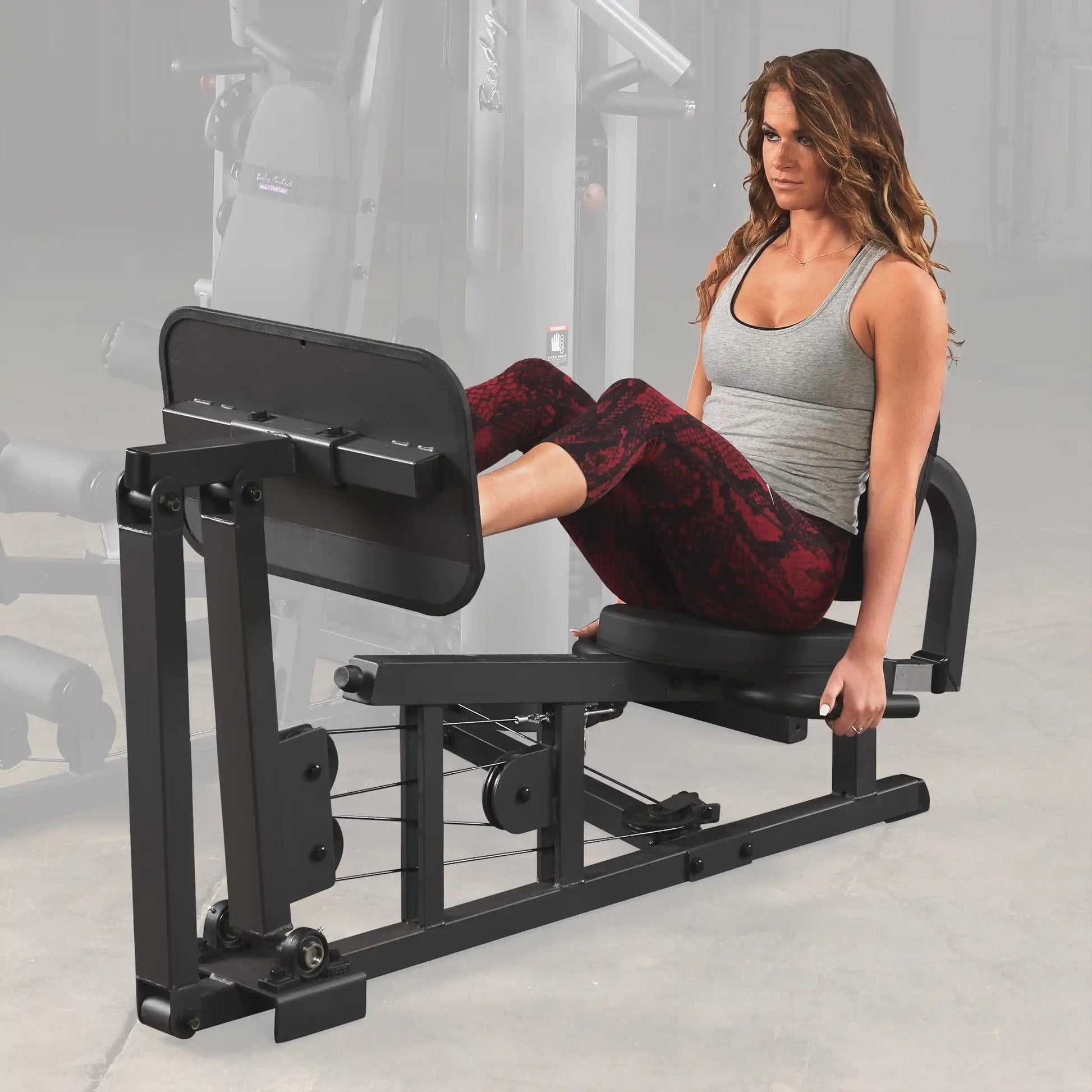 Woman using Body-Solid leg press machine in gym, wearing gray top and red leggings.