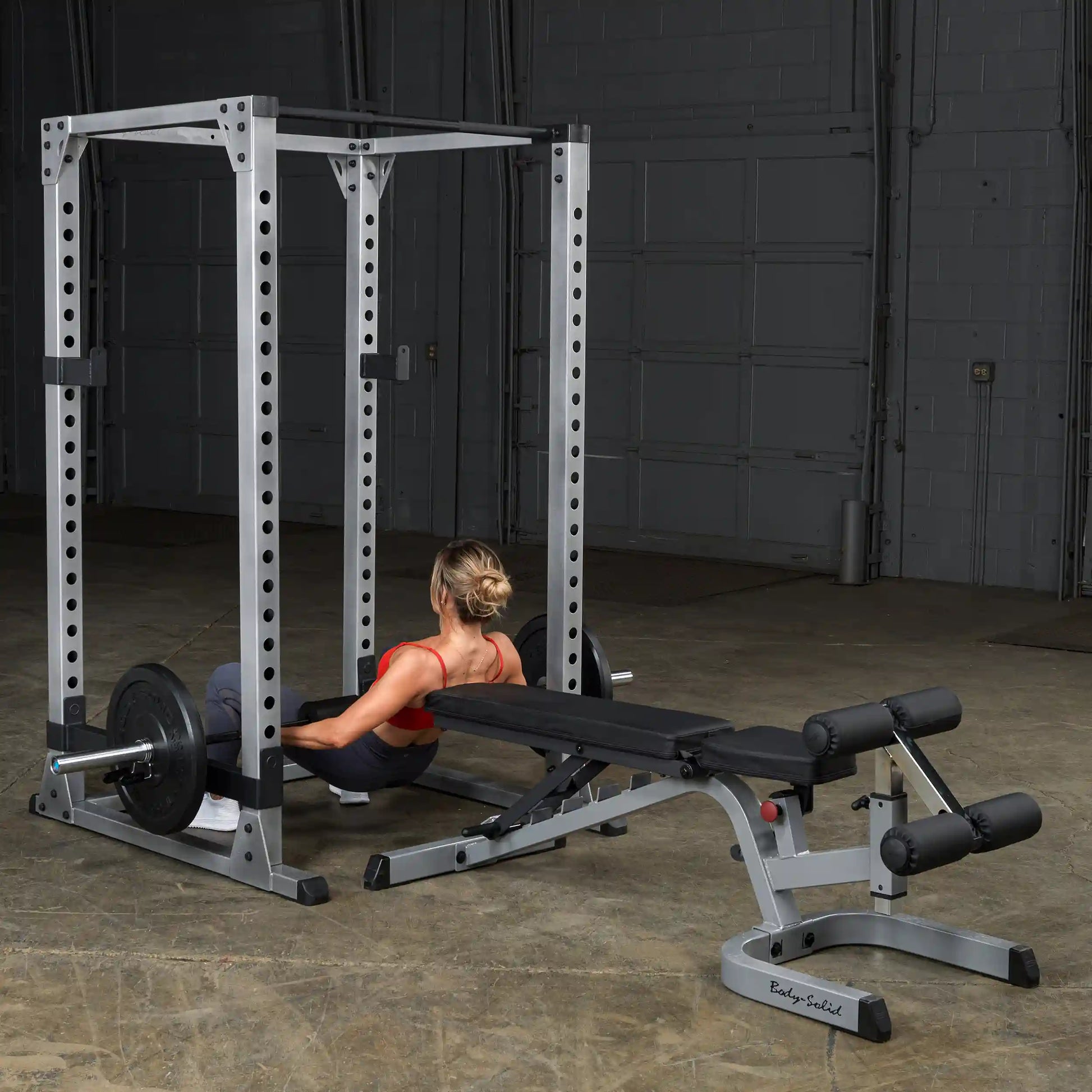 Woman using Body-Solid power rack with weight bench for strength training in gym.