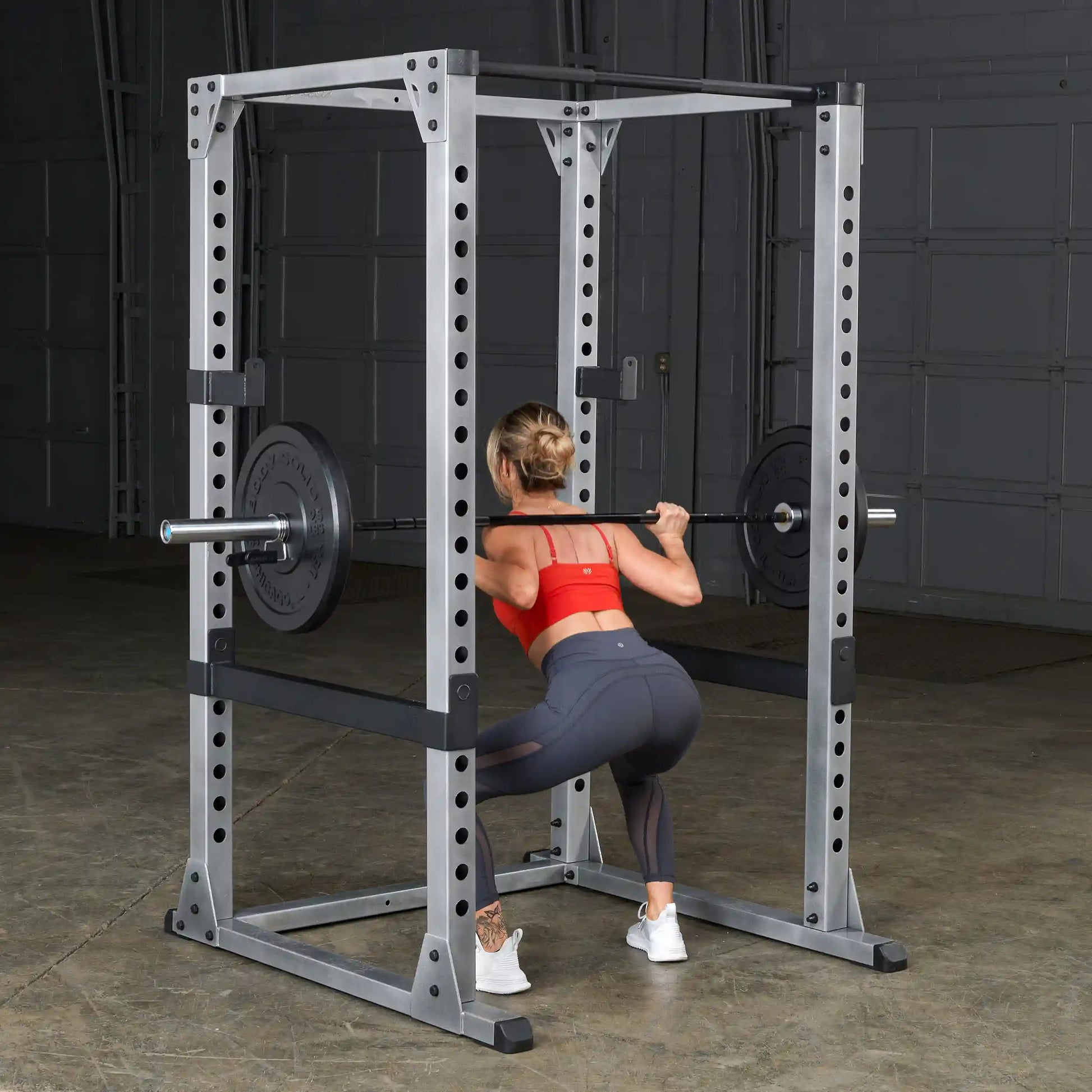 Woman performing squat in a power rack with barbell, wearing red sports bra and gray leggings.
