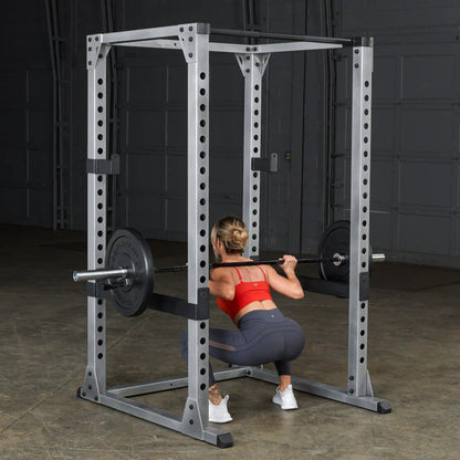 Woman squatting with barbell in a steel power rack, wearing a red sports bra and gray leggings.
