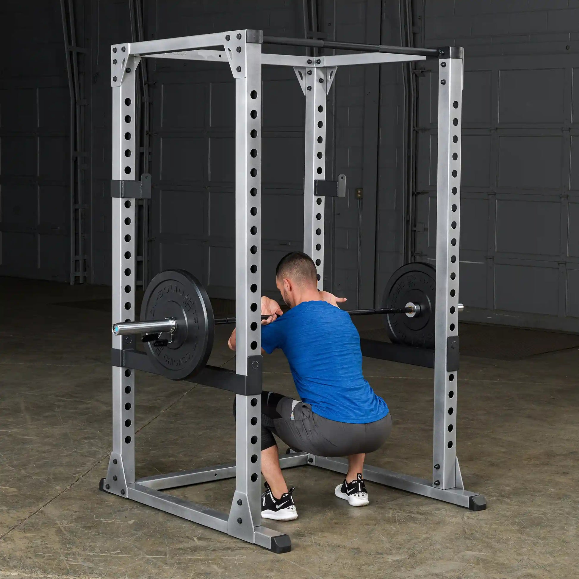 Man squatting inside a steel power rack with a barbell in a gym setting.