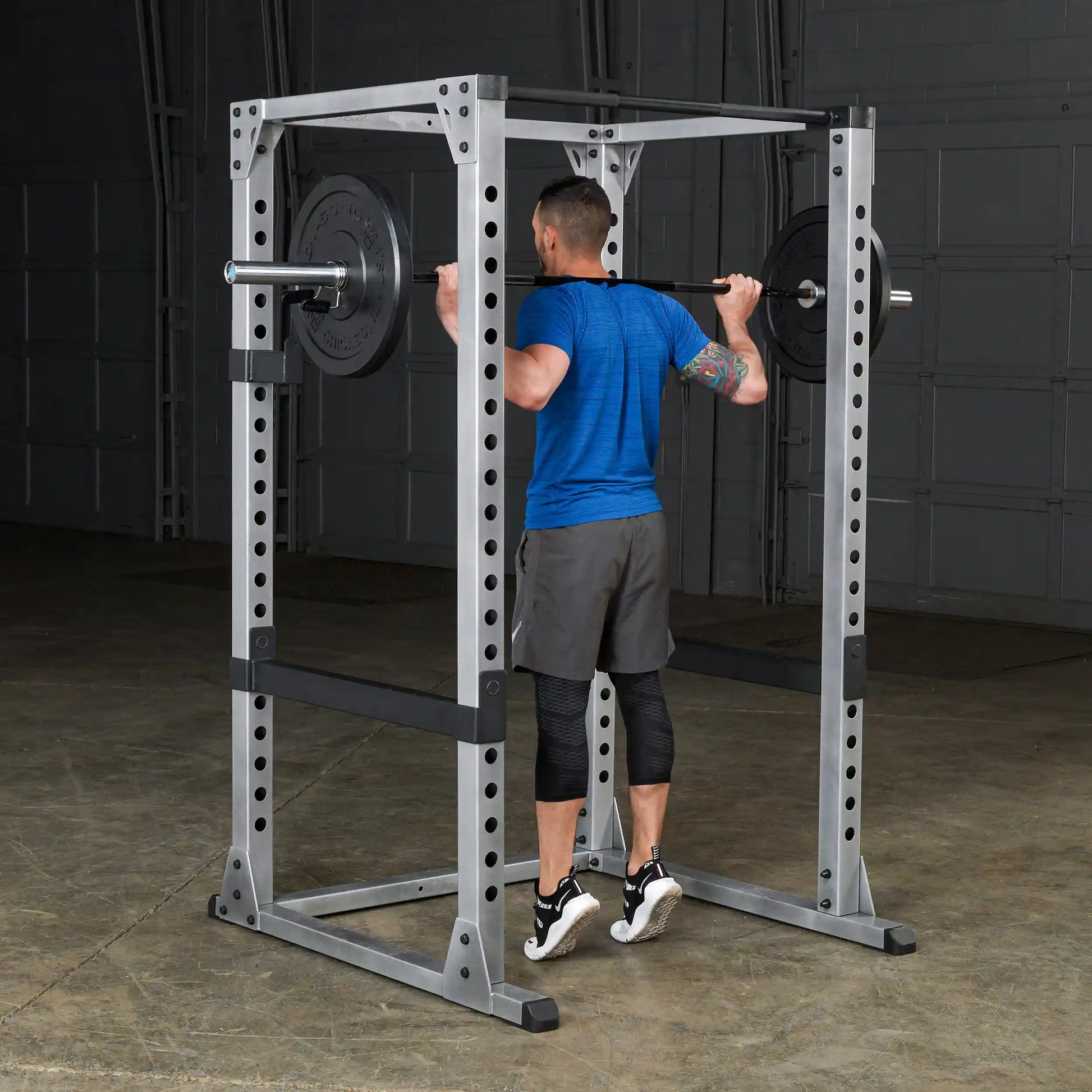 Man lifting weights on a gray power rack, wearing blue shirt and black shorts, performing a squat exercise.