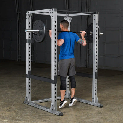 Man lifting weights on a gray power rack, wearing blue shirt and black shorts, performing a squat exercise.