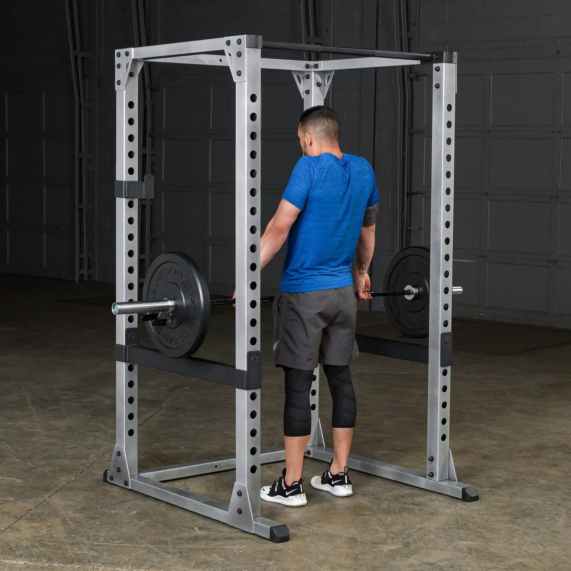 Man lifting weights in a Powerline squat rack, wearing a blue shirt and black knee sleeves.