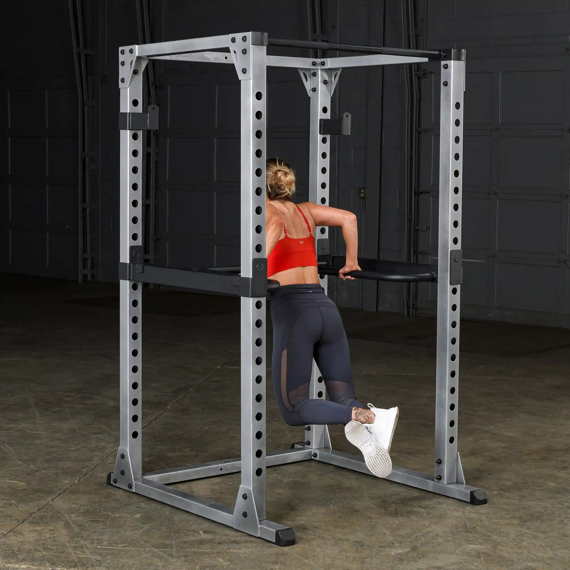 Woman using power rack for tricep dips in gym, wearing red sports bra and black leggings.