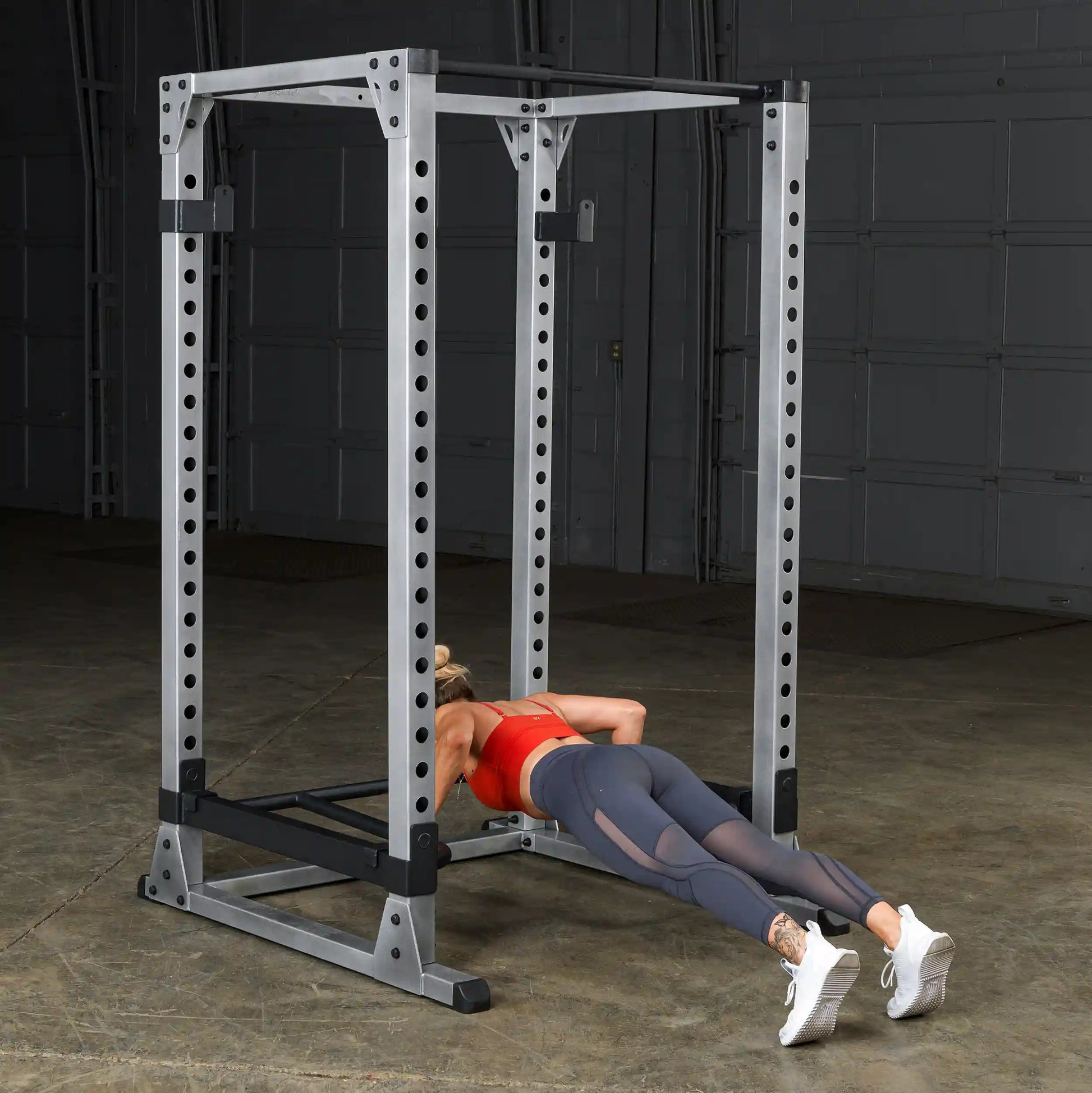Woman doing push-ups on a power rack in a gym, wearing a red top and gray leggings.