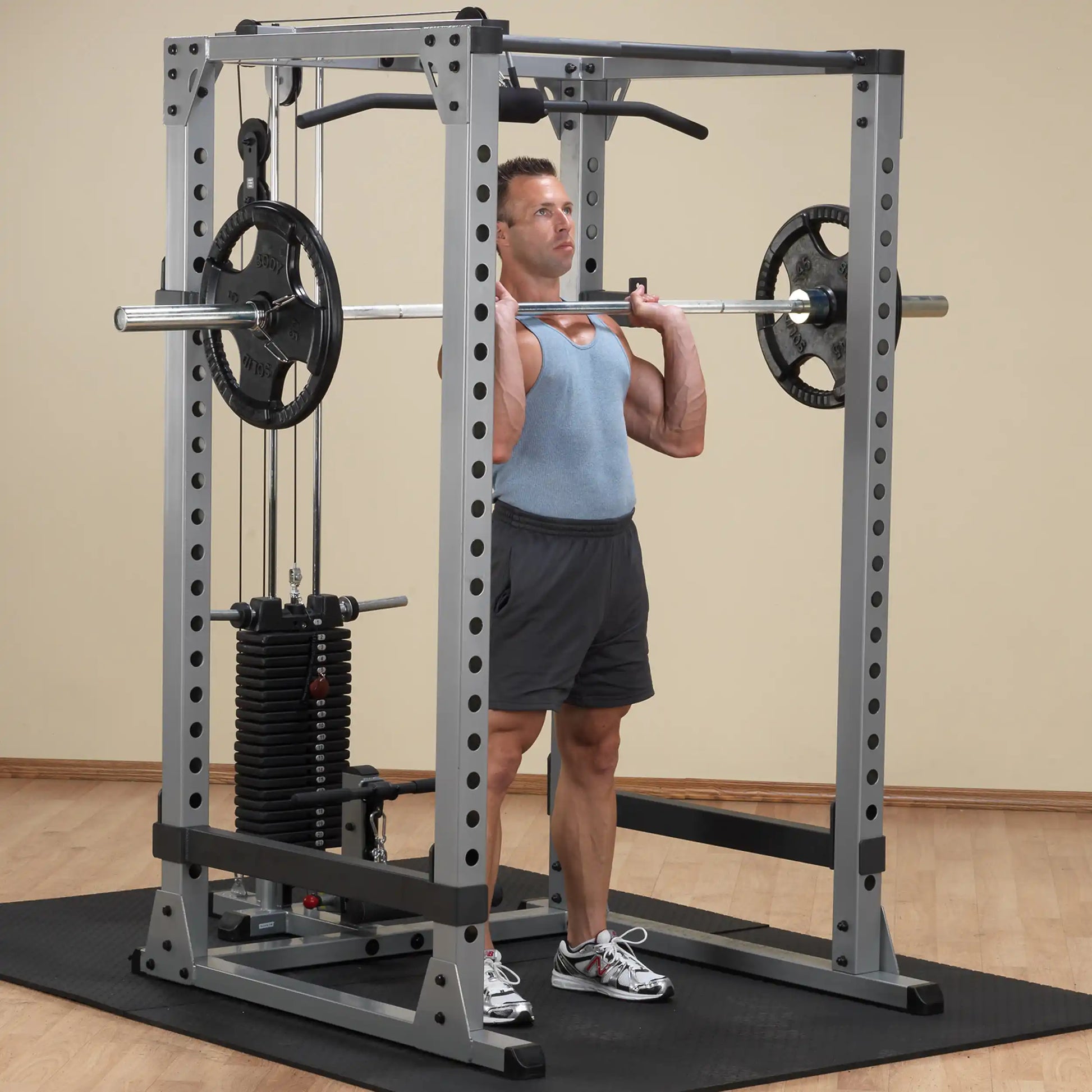 Man lifting barbell on Smith machine in gym, wearing blue tank top and gray shorts.