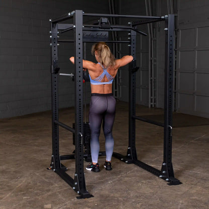 Woman using Body-Solid power rack in gym, wearing blue sports bra and black leggings.