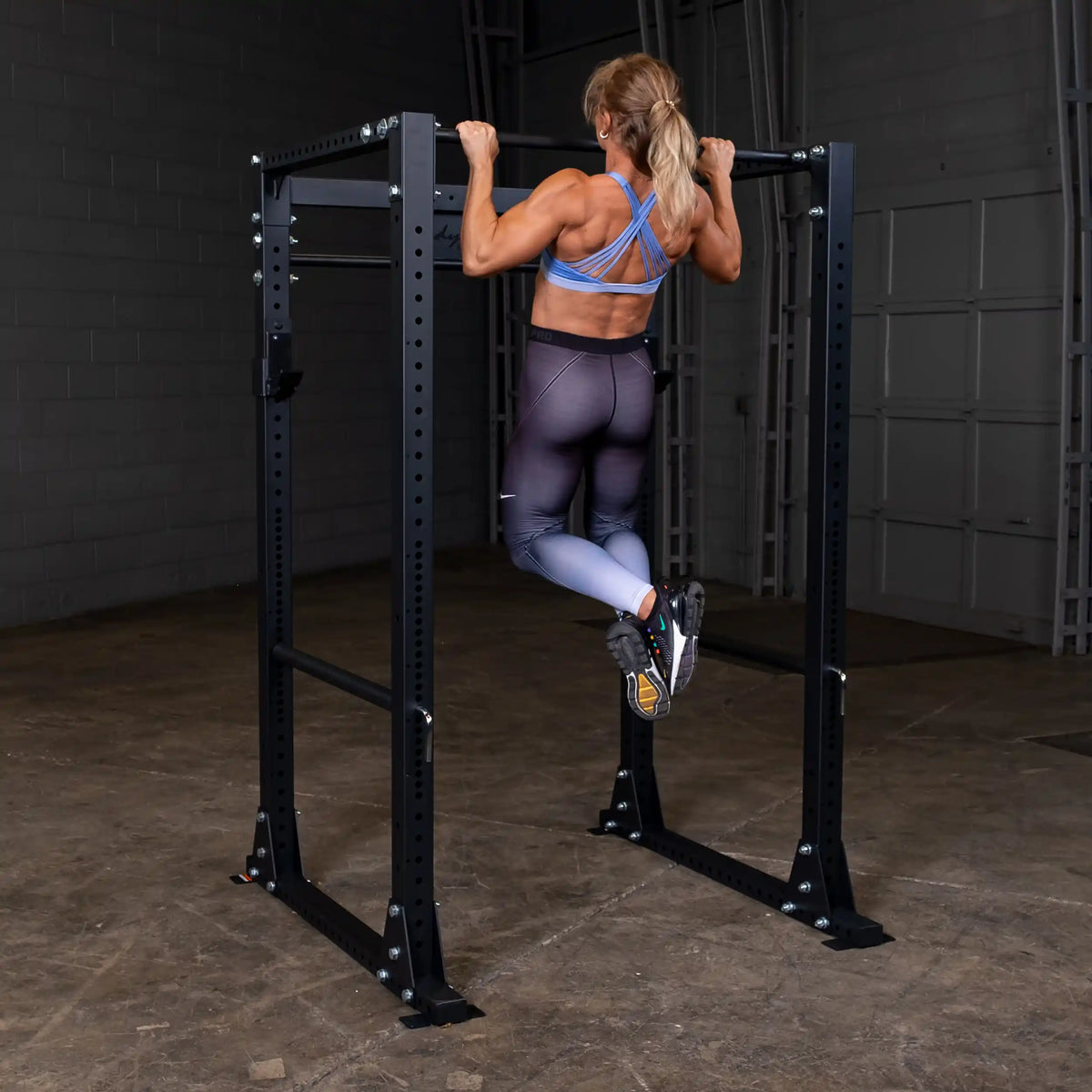 Woman doing pull-ups on black power rack in gym, wearing blue sports bra and gradient leggings.