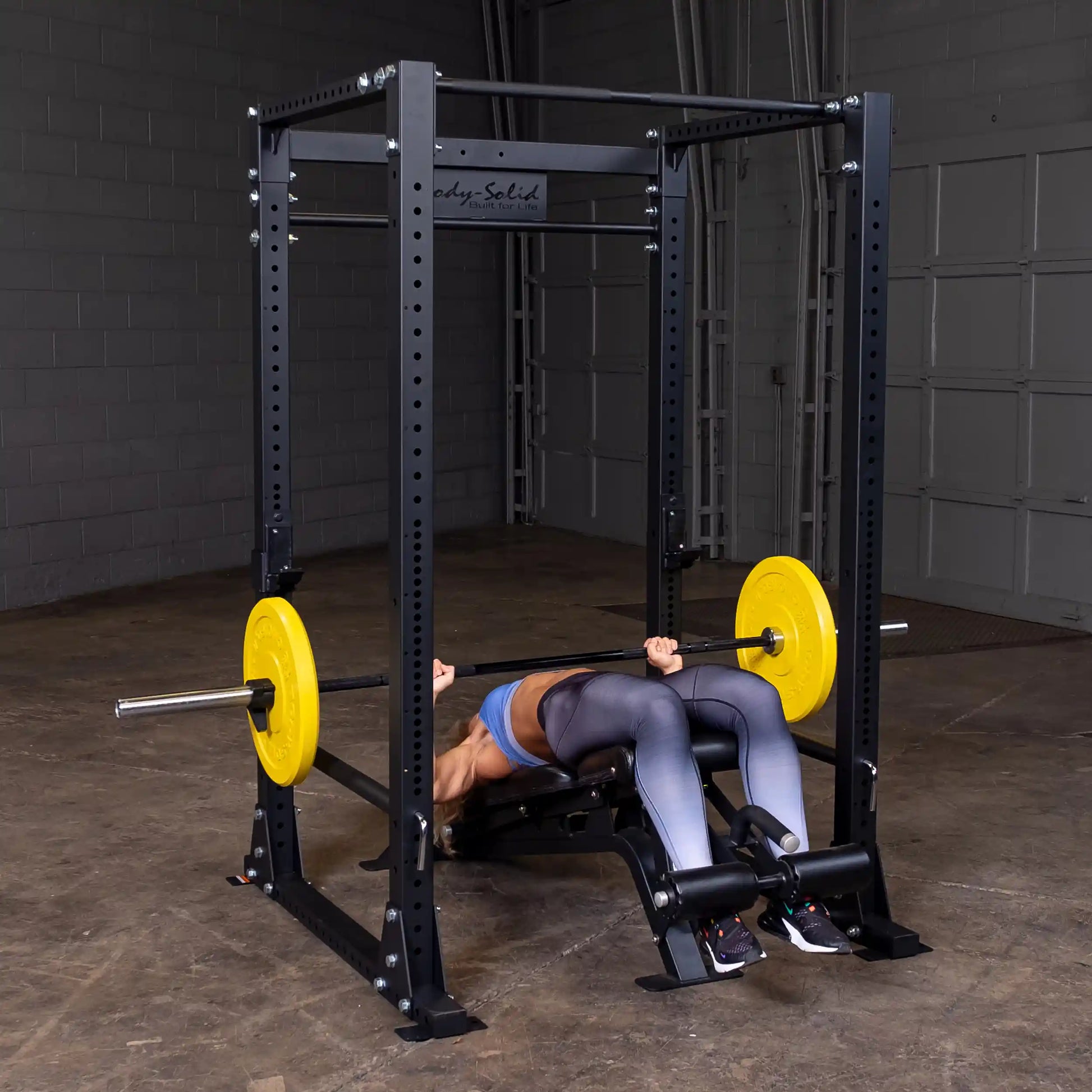 Person bench pressing on Body-Solid power rack with yellow weight plates in a gym.