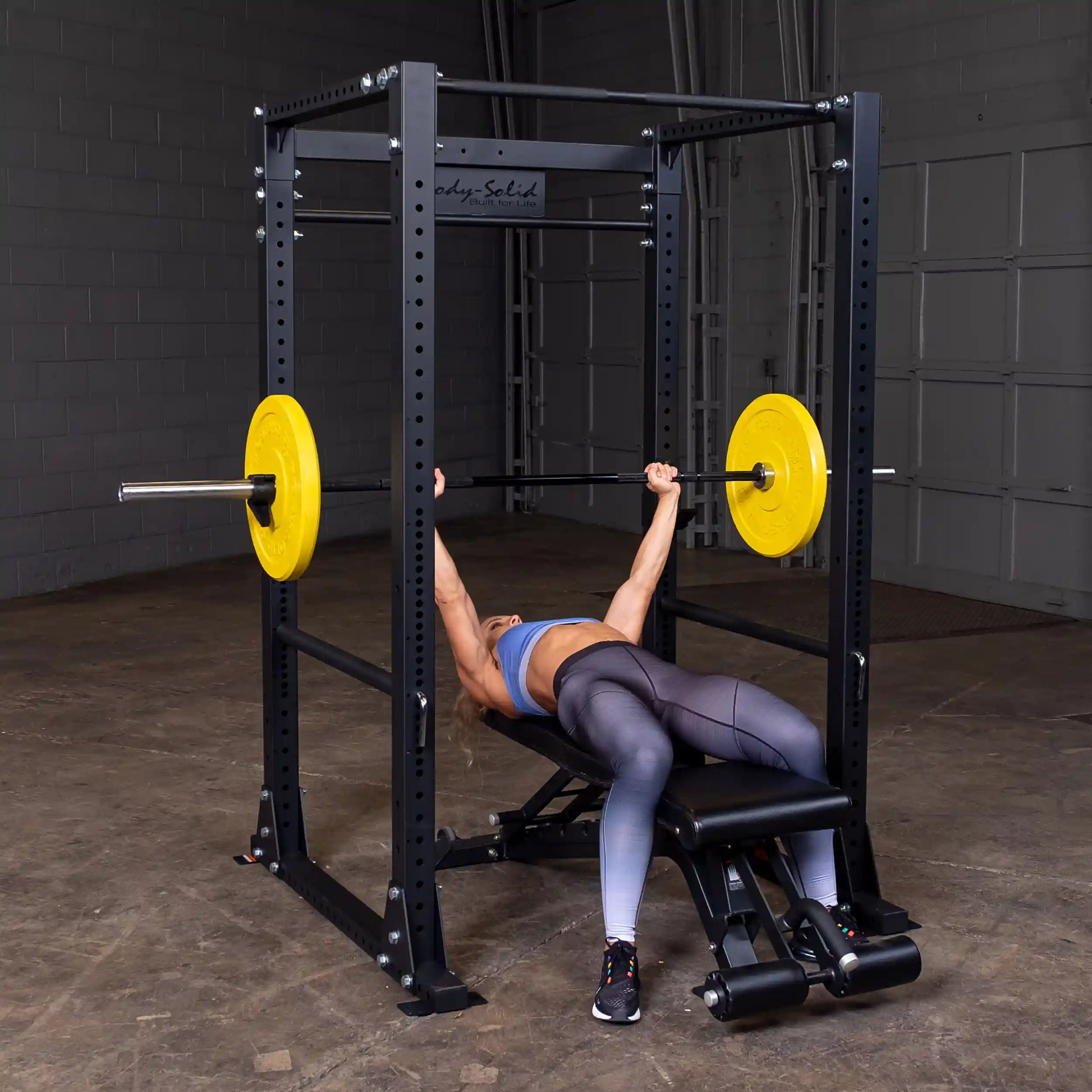 Woman bench pressing on Body-Solid power rack with yellow weight plates.
