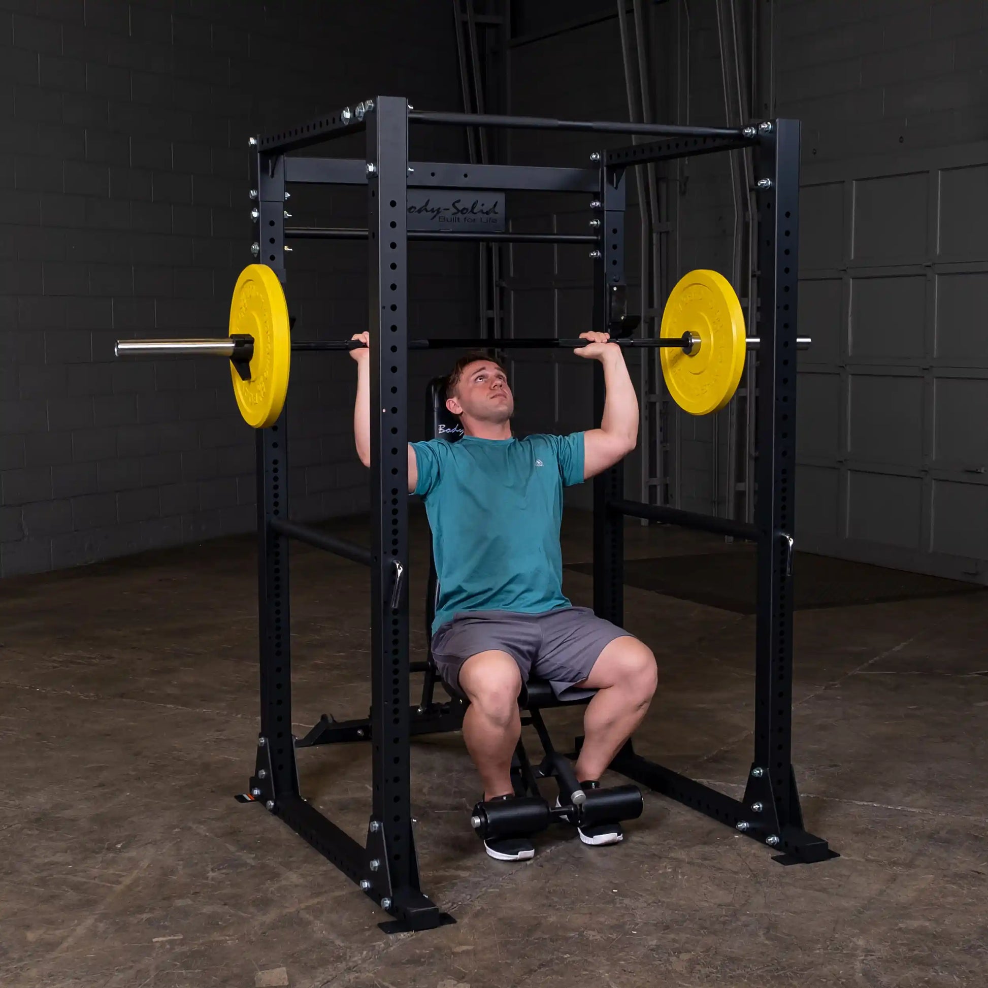 Man lifting barbell on Body-Solid power rack with yellow weights, wearing a teal shirt.