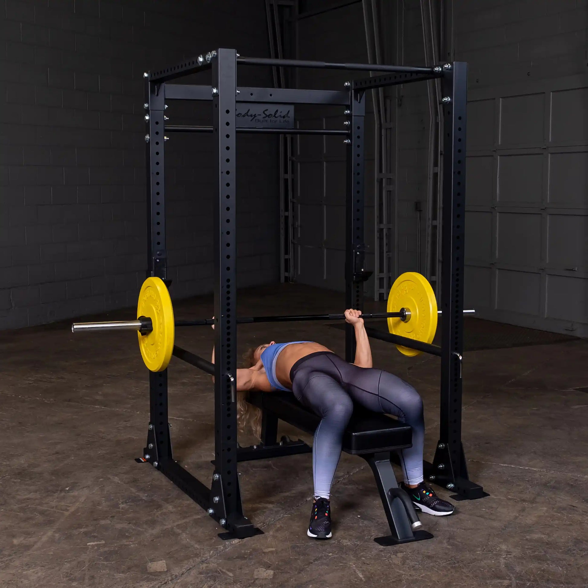 Woman bench pressing on Body-Solid power rack with yellow plates in a gym.