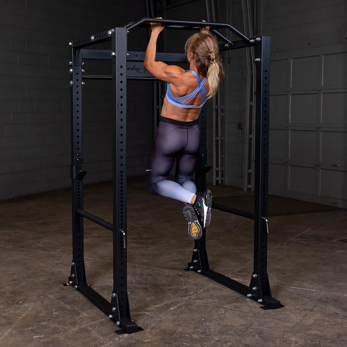 Woman doing pull-up on black power rack in gym.