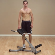 A shirtless man standing next to a workout bench, smiling, wearing black shorts and sneakers, in a gym setting with a wooden floor and beige walls.