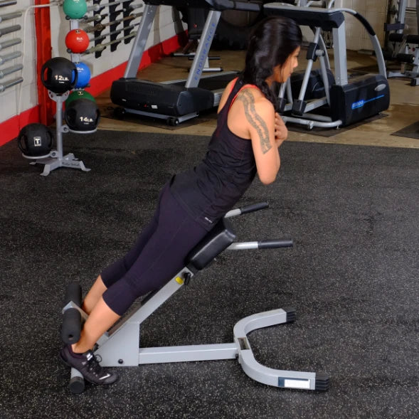 A person performing a back extension exercise on a gym machine, wearing a black tank top and leggings, with various gym equipment and weights visible in the background.
