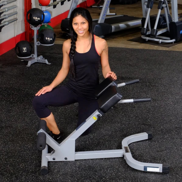 A woman in a black tank top and leggings kneels next to a gym equipment in a fitness center, smiling at the camera.
