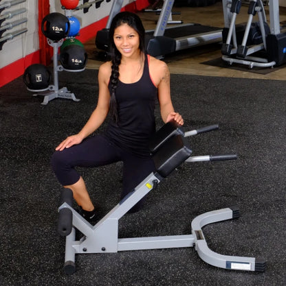 A woman in a black tank top and leggings kneels next to a gym equipment in a fitness center, smiling at the camera.