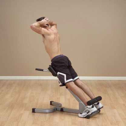 A shirtless man performing an abdominal exercise on a decline bench, with his hands clasped behind his head, in a fitness studio with a wooden floor and plain wall.
