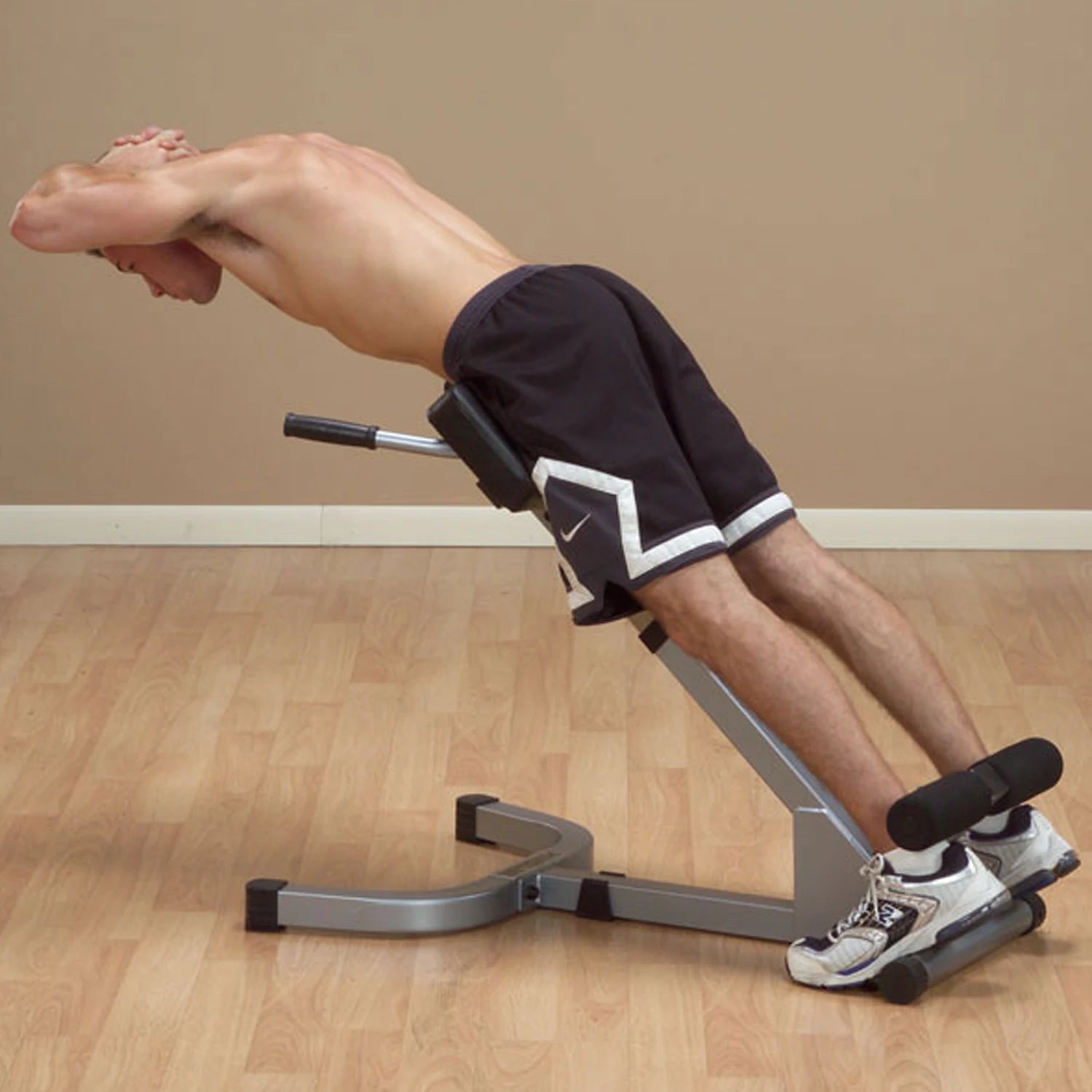A shirtless man performing a back extension exercise on a workout bench, leaning backwards with his hands behind his head, wearing black shorts and athletic shoes, in a fitness studio setting.