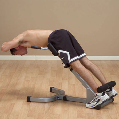 A shirtless man performing a back extension exercise on a fitness machine, leaning forward with his arms behind his head, demonstrating proper form.